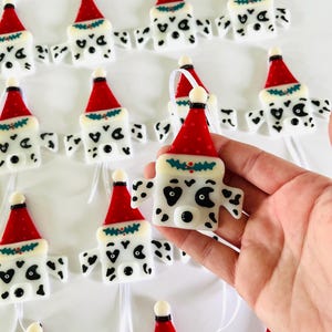 May include: A collection of handmade glass ornaments featuring a Dalmatian dog wearing a Santa hat. The ornaments are white with black spots and have a red Santa hat with a green holly sprig.