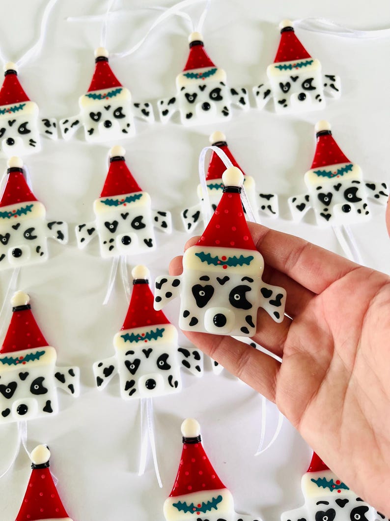 May include: A collection of handmade glass ornaments featuring a Dalmatian dog wearing a Santa hat. The ornaments are white with black spots and have a red Santa hat with a green holly sprig.