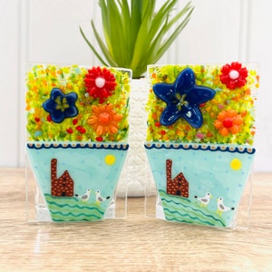 Könnte beinhalten: Zwei Glasmosaik-Blumentopf-Ornamente mit blauen, roten, orangen und gelben Blumen. Die Töpfe sind hellblau mit einem braunen Haus und zwei weißen Vögeln auf einer grünen Welle.