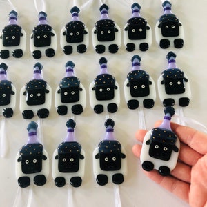 Puede incluir: Una colecci&oacute;n de 15 adornos de ovejas de vidrio blanco con caras negras y sombreros morados con lunares dorados. Cada oveja tiene una cinta blanca atada a la parte superior de su cabeza.