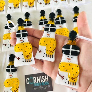 May include: A collection of handmade glass snowman ornaments with black hats, white bodies, and yellow and black patterned scarves. The ornaments are hanging from white ribbon and are displayed on a hand. The label reads "Cornish Fused Glass by Kirsty Smith".