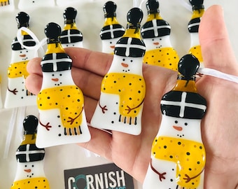 Puede incluir: Una colección de adornos de muñeco de nieve de vidrio hechos a mano con sombreros negros, cuerpos blancos y bufandas con patrones amarillos y negros. Los adornos cuelgan de una cinta blanca y se muestran en una mano. La etiqueta dice "Cornish Fused Glass by Kirsty Smith".