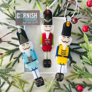 May include: Three fused glass Christmas tree decorations in the shape of toy soldiers. The ornaments are in red, blue, and yellow uniforms with black hats and boots. Each has a white ribbon for hanging. The ornaments are in a white box with a logo that says "Cornish Fused Glass".