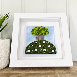 Könnte beinhalten: Ein gerahmtes Glaskunstwerk, das einen grünen Baum auf einem grünen Hügel mit weißen Schafen und roten Blumen darstellt. Der Rahmen ist weiß.