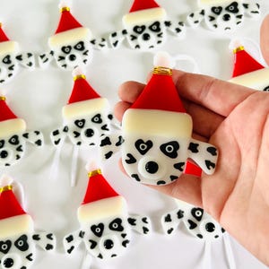 May include: A collection of handmade glass ornaments featuring Dalmatian dog faces wearing Santa hats. The ornaments are white with black spots and have black eyes and noses. Each ornament has a white ribbon for hanging.