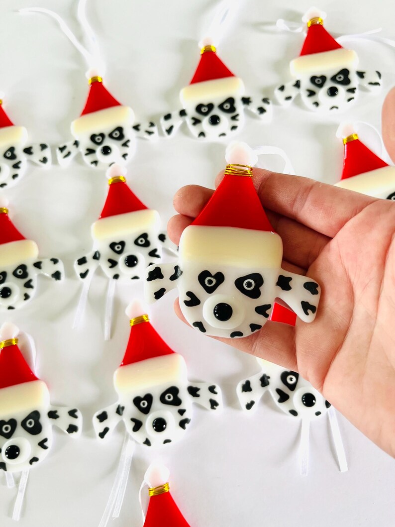 May include: A collection of handmade glass ornaments featuring Dalmatian dog faces wearing Santa hats. The ornaments are white with black spots and have black eyes and noses. Each ornament has a white ribbon for hanging.