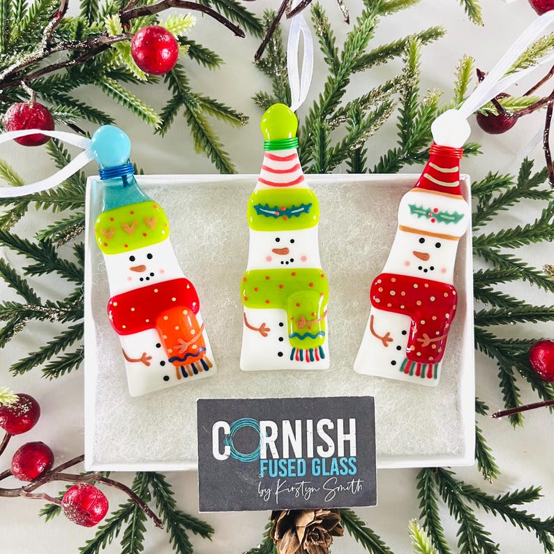 K&ouml;nnte beinhalten: Drei schneemannf&ouml;rmige Weihnachtsschmuckst&uuml;cke mit wei&szlig;en B&auml;ndern zum Aufh&auml;ngen. Jedes Ornament hat einen andersfarbigen Hut und Schal. Die Ornamente befinden sich in einer wei&szlig;en Schachtel, umgeben von Tannenzweigen und roten Beeren. Hergestellt aus verschmolzenem Glas.