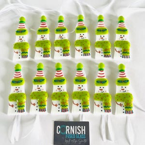 K&ouml;nnte beinhalten: Glas-Schneemann-Ornamente. Jeder Schneemann hat einen wei&szlig;en K&ouml;rper, einen gr&uuml;nen Schal mit roten Punkten und einen rot-wei&szlig; gestreiften Hut. Die Ornamente sind auf einer wei&szlig;en Oberfl&auml;che angeordnet.