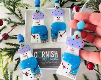 Puede incluir: Cuatro adornos de vidrio soplado hechos a mano con detalles en azul y blanco. Cada muñeco de nieve lleva una bufanda azul y un gorro morado con lunares blancos. Los adornos están decorados con pintura dorada y tienen un pequeño pájaro rojo en el gorro. Los adornos están etiquetados como "Cornish Fused Glass by Kirsty Smith".