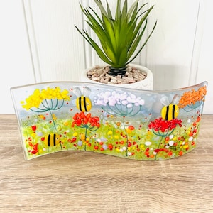 May include: A curved glass art piece with a whimsical design featuring yellow, orange, and red flowers with bees flying around them. The background is a light blue with white dots.