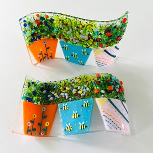 May include: A glass art piece featuring four colourful flower pots with different designs. The pots are arranged in a wave pattern on a clear glass background. The designs include bees, flowers, and geometric patterns.