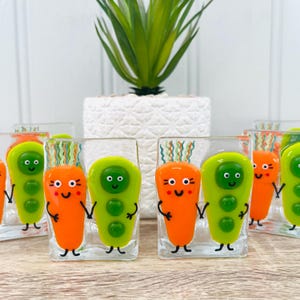 May include: A collection of clear glass shot glasses with cartoon characters in orange and green. Each glass features a smiling character with black arms and legs. The glasses are displayed on a wooden surface, with a white pot and plant in the background.