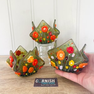 May include: Three fused glass bowls with a floral design. The bowls are olive green with orange, red, and yellow flowers. The bowls have a square shape with curved edges. The text "CORNISH FUSED GLASS by Kirsty Smith" is on a white rectangle.