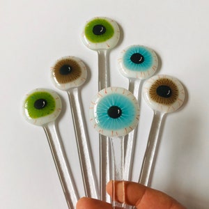 Puede incluir: Seis agitadores de bebidas de vidrio en forma de ojo con iris de diferentes colores. Los agitadores son de vidrio transparente con fondo blanco y venas rojas. Los iris son verdes, marrones, azules y negros.