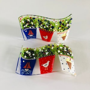May include: A set of two glass art pieces featuring a row of three flower pots with different designs. The pots are painted in red, white, and blue with seagull, sailboat, and ice cream cone designs. The flower pots are set against a background of green and white glass.