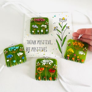 Puede incluir: Cuatro adornos cuadrados de vidrio con fondo verde, que representan una flor blanca con abejas amarillas y negras. Los adornos cuelgan de cintas blancas. El texto "Think positive, bee positive!" está impreso en una tarjeta blanca con tinta negra y puntos de colores.