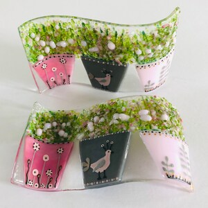 May include: Two glass mosaic wall hangings with a wavy design. Each piece features three potted plants with pink, grey, and white pots. The pots have a floral design and a small bird illustration.