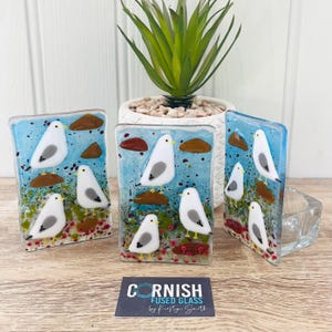 Könnte beinhalten: Vier kleine Glasscheiben mit blauem Hintergrund und weißen Möwen. Die Möwen sitzen auf braunen Felsen und im Vordergrund befinden sich rote und orangefarbene Blumen. Die Tafeln werden von Cornish Fused Glass von Kirsty Smith hergestellt.