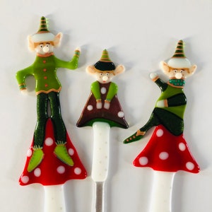 Könnte beinhalten: Drei gläserne Ornamente in Form von Elfen, die auf pilzförmigen Sockeln stehen. Die Elfen tragen grüne und braune Outfits mit weißen Akzenten. Die Pilze sind rot mit weißen Punkten.