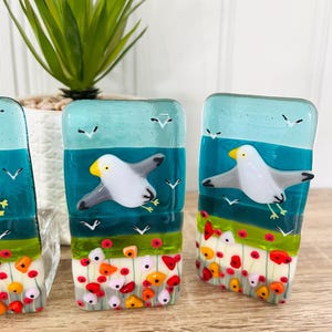 Könnte beinhalten: Rechteckige Glaskunststücke mit einem Möwen-Design. Das Kunstwerk zeigt einen türkisblauen Himmel, einen dunkel türkisblauen Ozean und ein Feld aus roten, orangen und weißen Blumen. Die Möwe hat einen gelben Schnabel und graue Flügel.