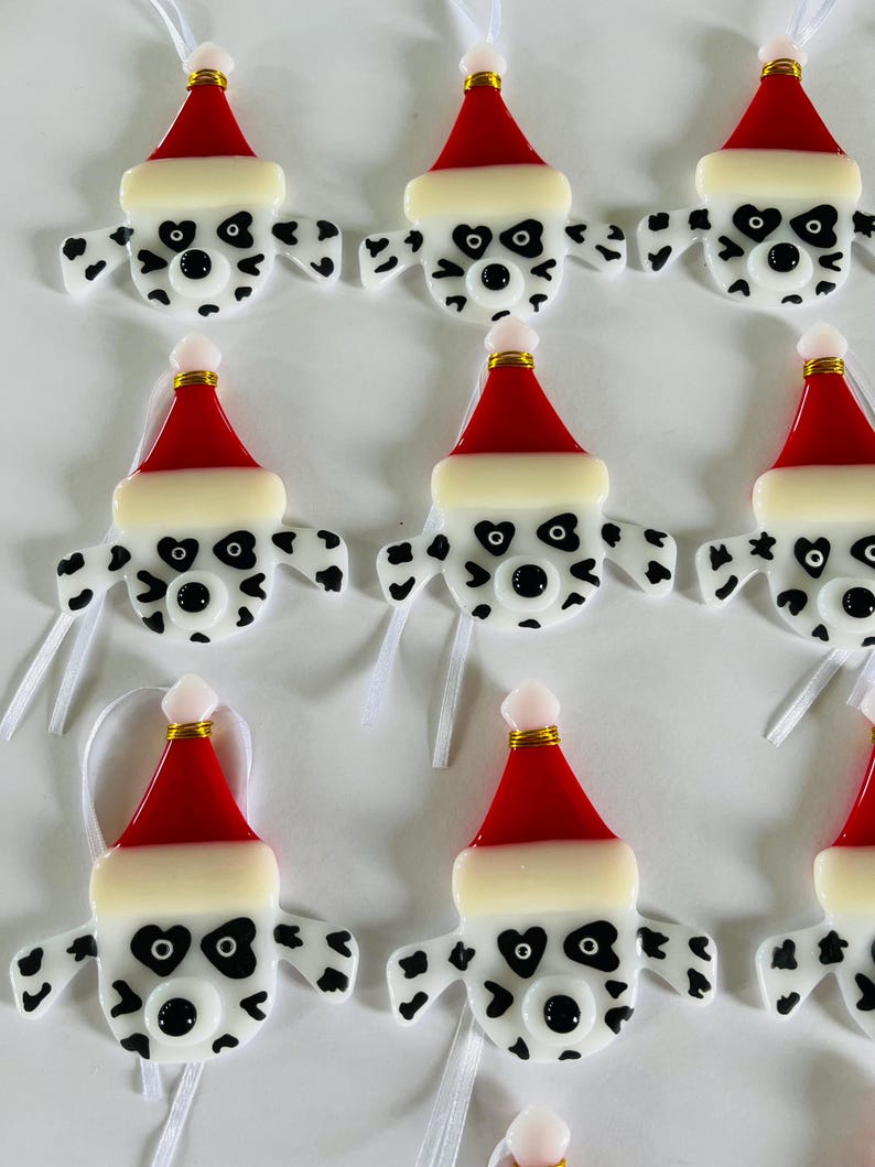 May include: Ten handmade glass ornaments featuring Dalmatian dog faces wearing Santa hats. The ornaments are white with black spots and have red and white Santa hats.