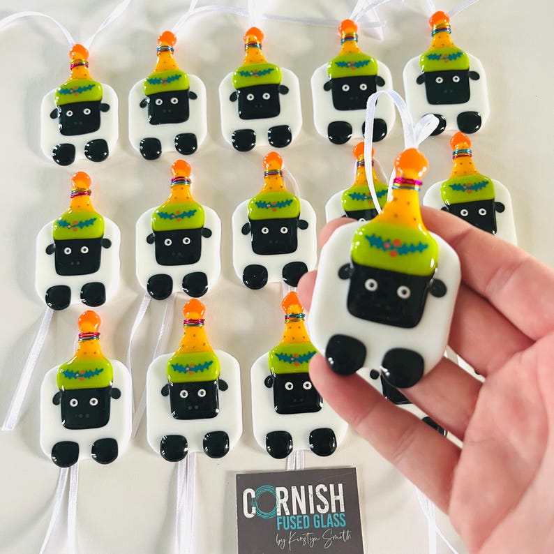 Puede incluir: Una colecci&oacute;n de 12 adornos de oveja de vidrio fundido blanco con ojos negros y sombreros de fiesta naranja y verde. Los adornos est&aacute;n colgados de cintas blancas y est&aacute;n dispuestos en filas. El texto "CORNISH FUSED GLASS by Kirstie Smith" es visible en la esquina inferior derecha de la imagen.