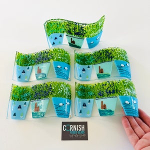 May include: Four pieces of fused glass art depicting a row of blue and white flower pots with various designs, including sailboats, sheep, and houses. The glass pieces are arranged in a wave pattern and are set against a white background. The artwork is signed "Cornish Fused Glass by Katie Smith".