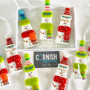 K&ouml;nnte beinhalten: Handgefertigte Schneemann-Ornamente aus verschmolzenem Glas. Jedes Ornament ist wei&szlig; mit einem bunten Schal, Hut und Kn&ouml;pfen. Die Ornamente werden in einer wei&szlig;en Schachtel pr&auml;sentiert und sind fertig zum Aufh&auml;ngen. Die Ornamente sind etwa 10 cm hoch.