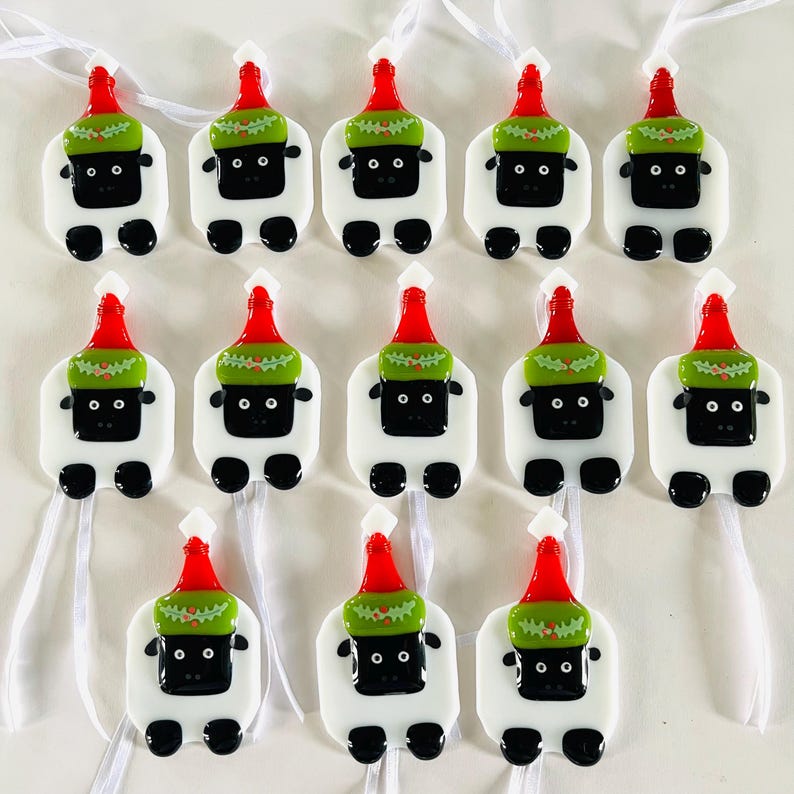 Puede incluir: Doce adornos de vidrio blanco con forma de oveja que llevan gorros de Pap&aacute; Noel rojos y verdes. Los adornos tienen ojos negros y una nariz negra. Cada adorno tiene una cinta blanca.