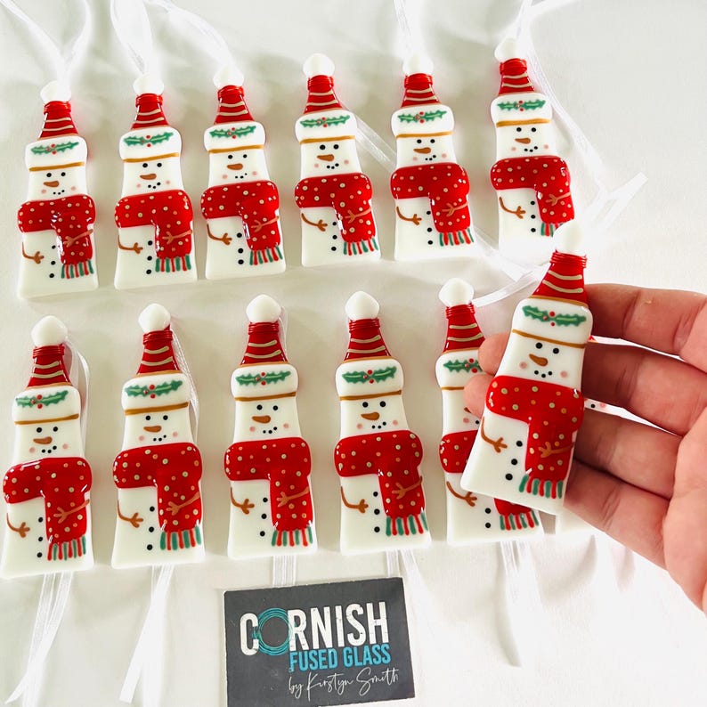 K&ouml;nnte beinhalten: Eine Sammlung von schneemannf&ouml;rmigen Weihnachtsschmuck. Jedes Ornament ist wei&szlig; mit rotem Hut, Schal und Akzenten. Die Ornamente sind mit wei&szlig;en B&auml;ndern aufgereiht. Das Bild enth&auml;lt eine Visitenkarte mit der Aufschrift "CORNISH FUSED GLASS by Kirstyn Smith."