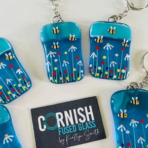 Peut inclure: Porte-clés en verre fusionné de forme rectangulaire, avec un dessus turquoise et une base bleue. La base présente un motif floral avec des fleurs blanches, des points rouges et jaunes, et de petites abeilles. Les porte-clés ont une chaîne et un anneau argentés.