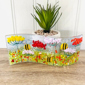 May include: A glass art piece depicting a field of flowers with bees. The flowers are yellow, red, and white. The bees are yellow and black. The background is a light blue.