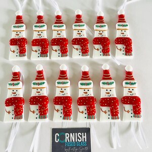 K&ouml;nnte beinhalten: Eine Sammlung von schneemannf&ouml;rmigen Weihnachtsschmuck. Jeder Schmuck ist wei&szlig; mit einem roten Schal und Hut sowie schwarzen Punktaugen und Mund. Die Ornamente sind mit wei&szlig;en B&auml;ndern aufgereiht. Das Bild enth&auml;lt ein schwarzes Schild mit der Aufschrift "CORNISH FUSED GLASS by Kirstyn Smith."