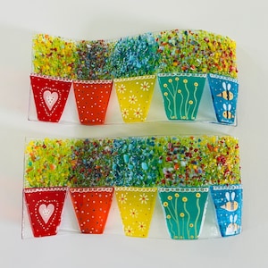 May include: Two rows of colourful glass flower pots with different floral designs. The pots are arranged in a wave pattern and are set against a white background.