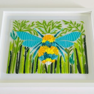 May include: A framed glass mosaic artwork depicting a bumblebee with yellow and blue wings, surrounded by green foliage.