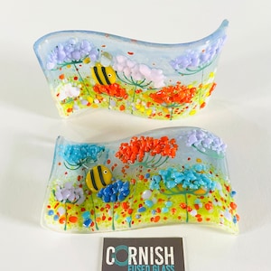 May include: Two wavy glass pieces with a blue background and a colourful floral design. The flowers are orange, pink, and blue with yellow bees. The glass pieces are textured with small dots and specks of colour. The bottom piece has the text "CORNISH FUSED GLASS".