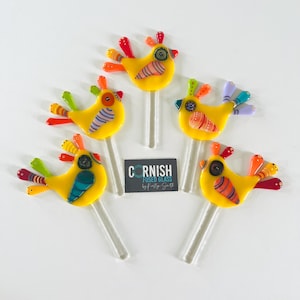 May include: Five yellow fused glass bird ornaments with colorful wings and tail feathers. Each bird has a clear glass stem. The birds have various patterns and colors, including red, orange, blue, and green. The text "CORNISH FUSED GLASS" is on a black and white card.