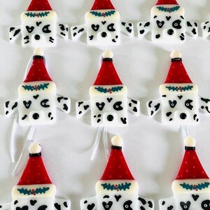 May include: Nine white glass ornaments shaped like Dalmatian dogs wearing red Santa hats with green holly trim. Each ornament has black spots and a black nose.