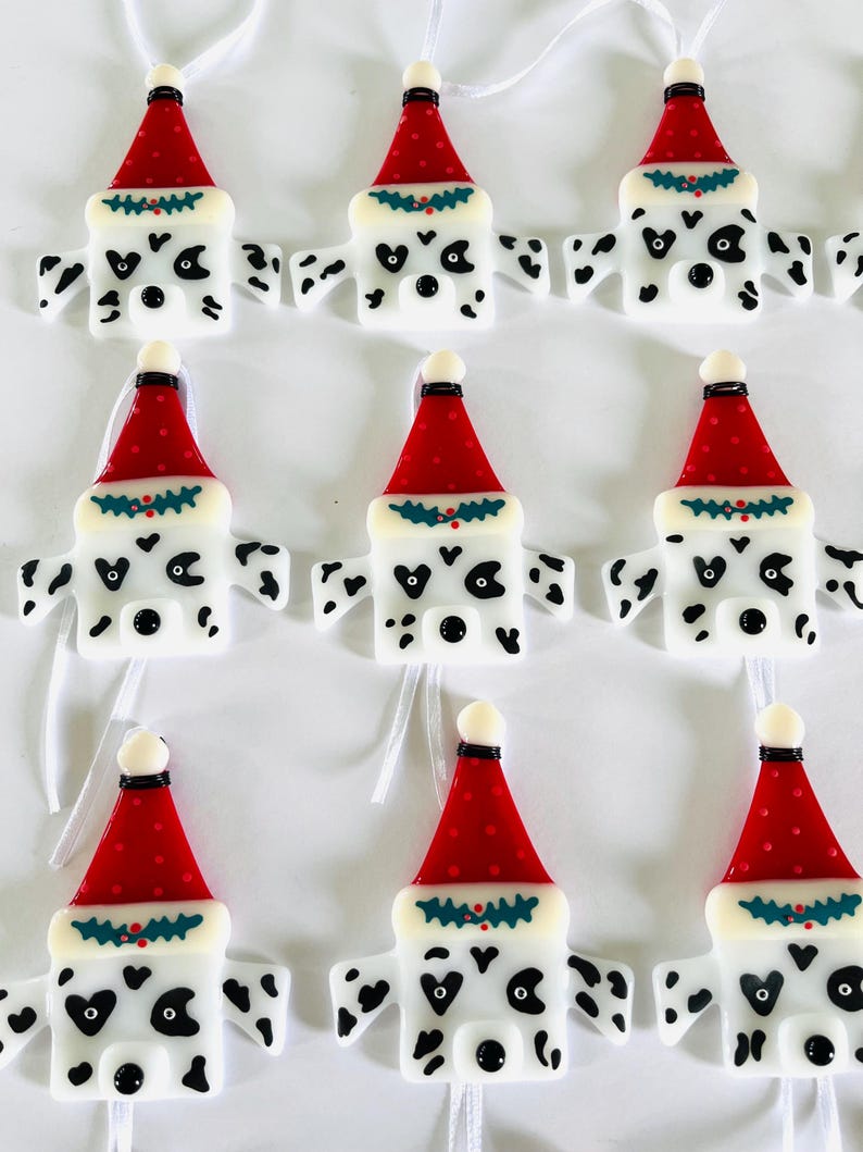May include: Nine white glass ornaments shaped like Dalmatian dogs wearing red Santa hats with green holly trim. Each ornament has black spots and a black nose.