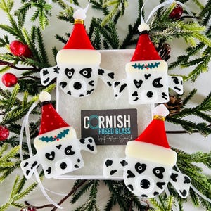 May include: Four handmade fused glass ornaments shaped like Dalmatian dogs wearing Santa hats. The ornaments are white with black spots and have black eyes and noses. The ornaments are hanging from a Christmas tree.