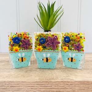 May include: Three small glass flower pots with a bee design. Each pot is filled with colourful glass flowers and beads. The pots are on a wooden surface.
