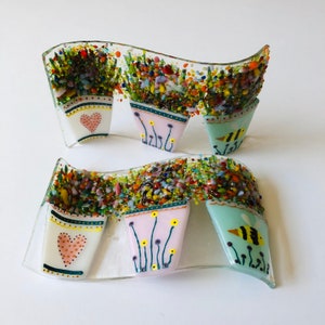 May include: A set of two glass mosaic wall art pieces featuring a row of flower pots with bees and hearts. The pots are decorated with colourful mosaic tiles and painted details.