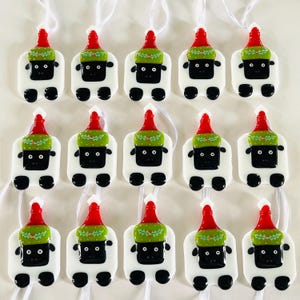 Puede incluir: Quince adornos de vidrio blanco con forma de oveja que llevan gorros de Pap&aacute; Noel rojos y verdes. Los adornos tienen ojos negros y una nariz negra. Cada adorno tiene una cuerda blanca para colgar.