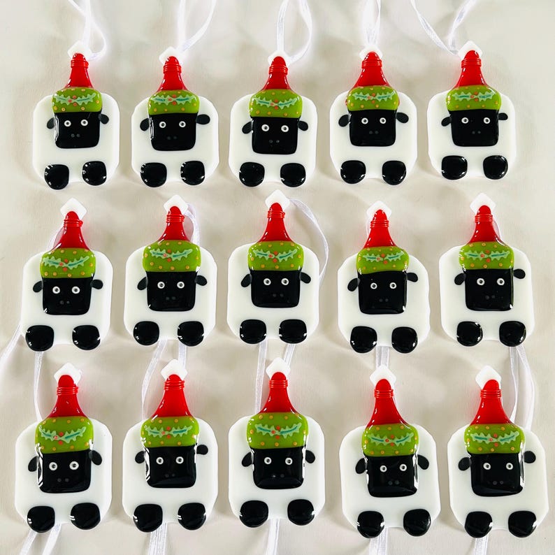 Puede incluir: Quince adornos de vidrio blanco con forma de oveja que llevan gorros de Pap&aacute; Noel rojos y verdes. Los adornos tienen ojos negros y una nariz negra. Cada adorno tiene una cuerda blanca para colgar.