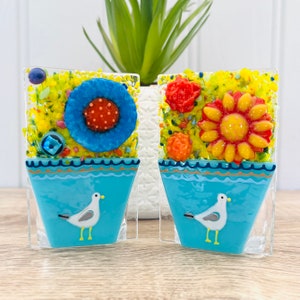 Könnte beinhalten: Zwei blaue Keramik-Blumentöpfe mit gelben, orangefarbenen und roten Blumen und einer weißen Möwe auf jedem Topf.