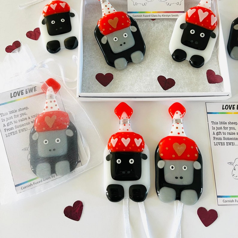 Puede incluir: Dos adornos de vidrio de ovejas blancas y negras con sombreros rojos y blancos. Las ovejas llevan sombreros de fiesta con lunares blancos y corazones rojos. El texto "LOVE EWE" est&aacute; en la tarjeta.