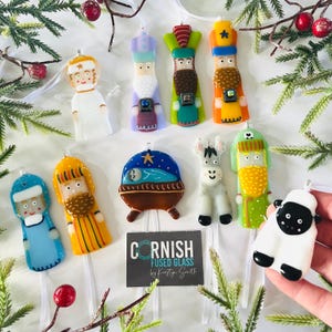 May include: A set of 10 fused glass ornaments depicting the Nativity scene. The ornaments are colorful and feature characters like the three wise men, a donkey, a sheep, and a baby Jesus in a manger. The ornaments are labeled "Cornish Fused Glass by Kirsty Smith".