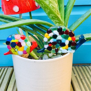 Pode incluir: Duas estacas de jardim de caracol de mosaico de vidro colorido em um vaso branco com uma planta de aloe verde e uma estaca de jardim de cogumelo vermelho e branco.