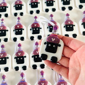 Puede incluir: Una colecci&oacute;n de adornos de vidrio hechos a mano con forma de oveja con sombreros morados y dorados. Los adornos tienen ojos negros y una cara negra. Los adornos cuelgan de una cinta blanca.
