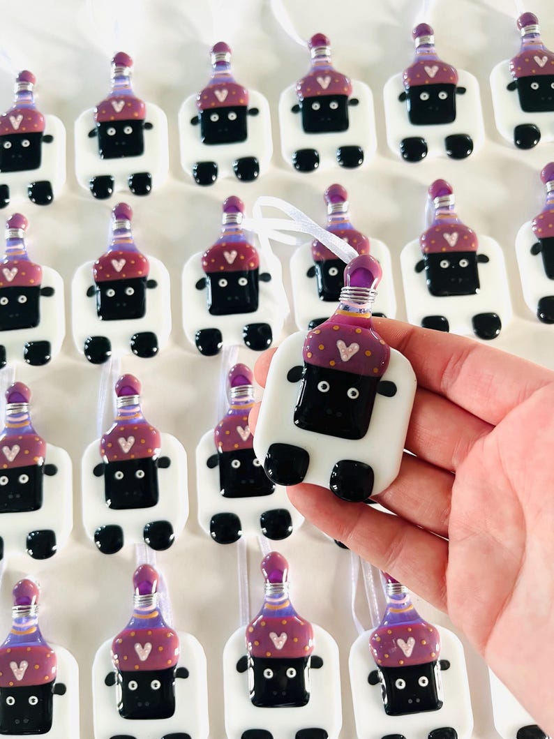 Puede incluir: Una colecci&oacute;n de adornos de vidrio hechos a mano con forma de oveja con sombreros morados y dorados. Los adornos tienen ojos negros y una cara negra. Los adornos cuelgan de una cinta blanca.